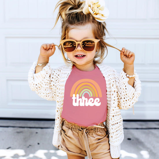 Flat lay of a purple toddler girls retro rainbow third birthday graphic tee