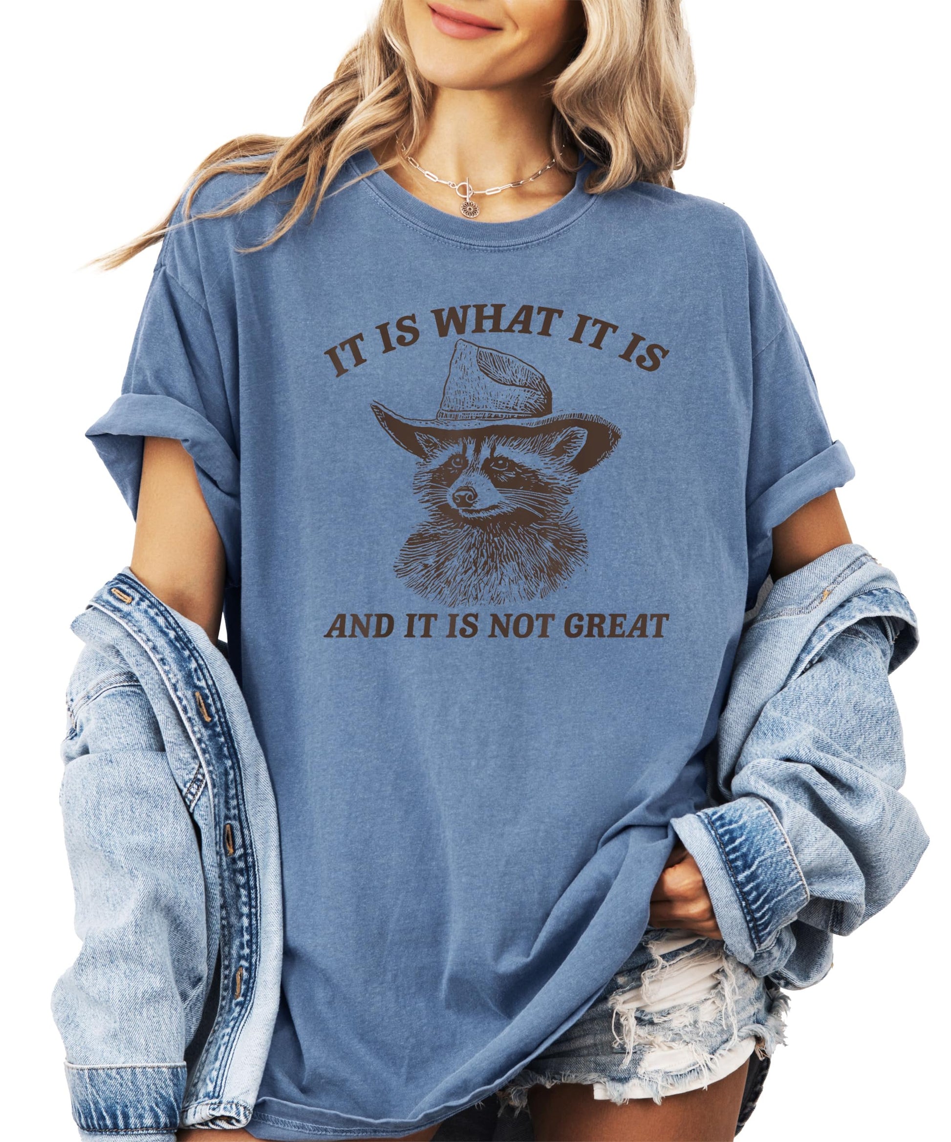 Ivory women's graphic tee with a cowboy raccoon and the phrase It Is What It Is and It Is Not Great printed on the front