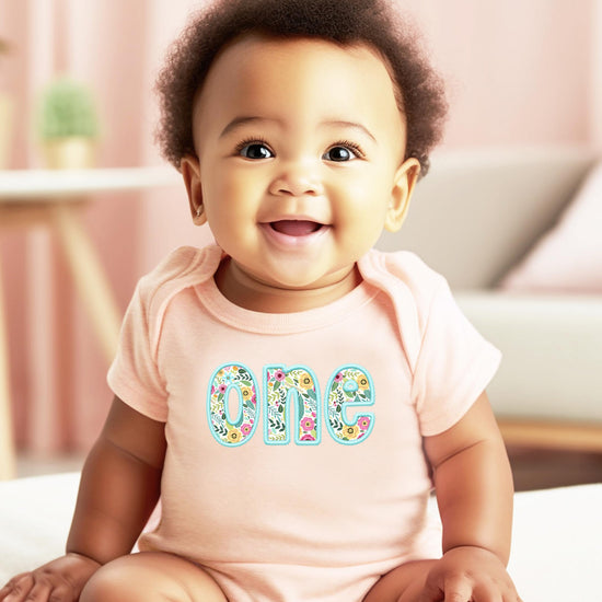 Close-up of teal embroidered floral lettering on peach 1st birthday baby girl bodysuit