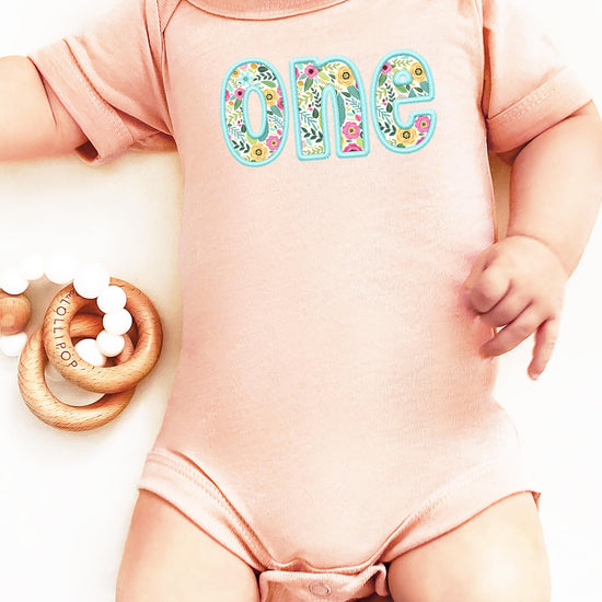 Flat lay of peach baby bodysuit with teal-trimmed floral first birthday embroidery print