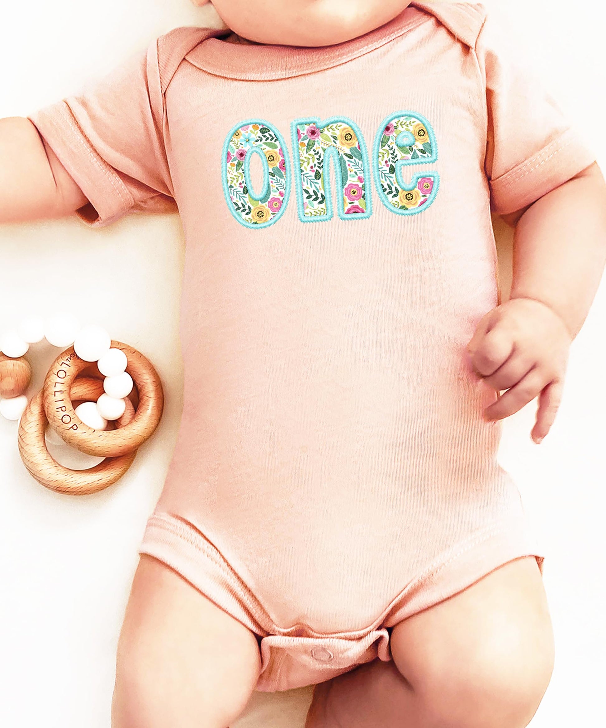Flat lay of peach baby bodysuit with teal-trimmed floral first birthday embroidery print