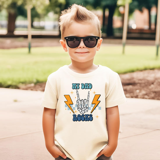 Front view of My Dad Rocks retro rocker skeleton hand kids t-shirt in natural
