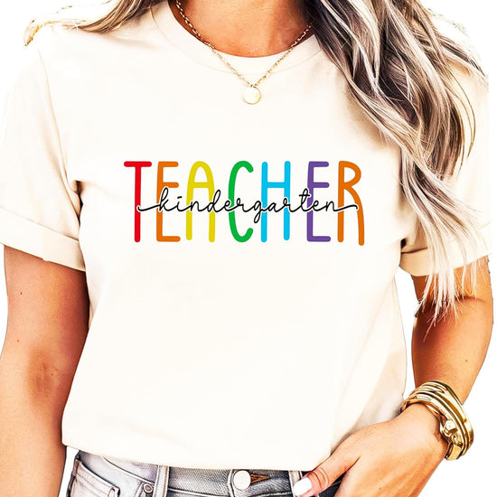 Women's pink kindergarten teacher shirt with bold rainbow TEACHER letters and hand-lettered script overlay