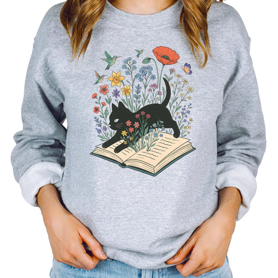 White sweatshirt with black cat sitting on open book surrounded by colorful flowers, birds, and butterflies — bookworm graphic design
