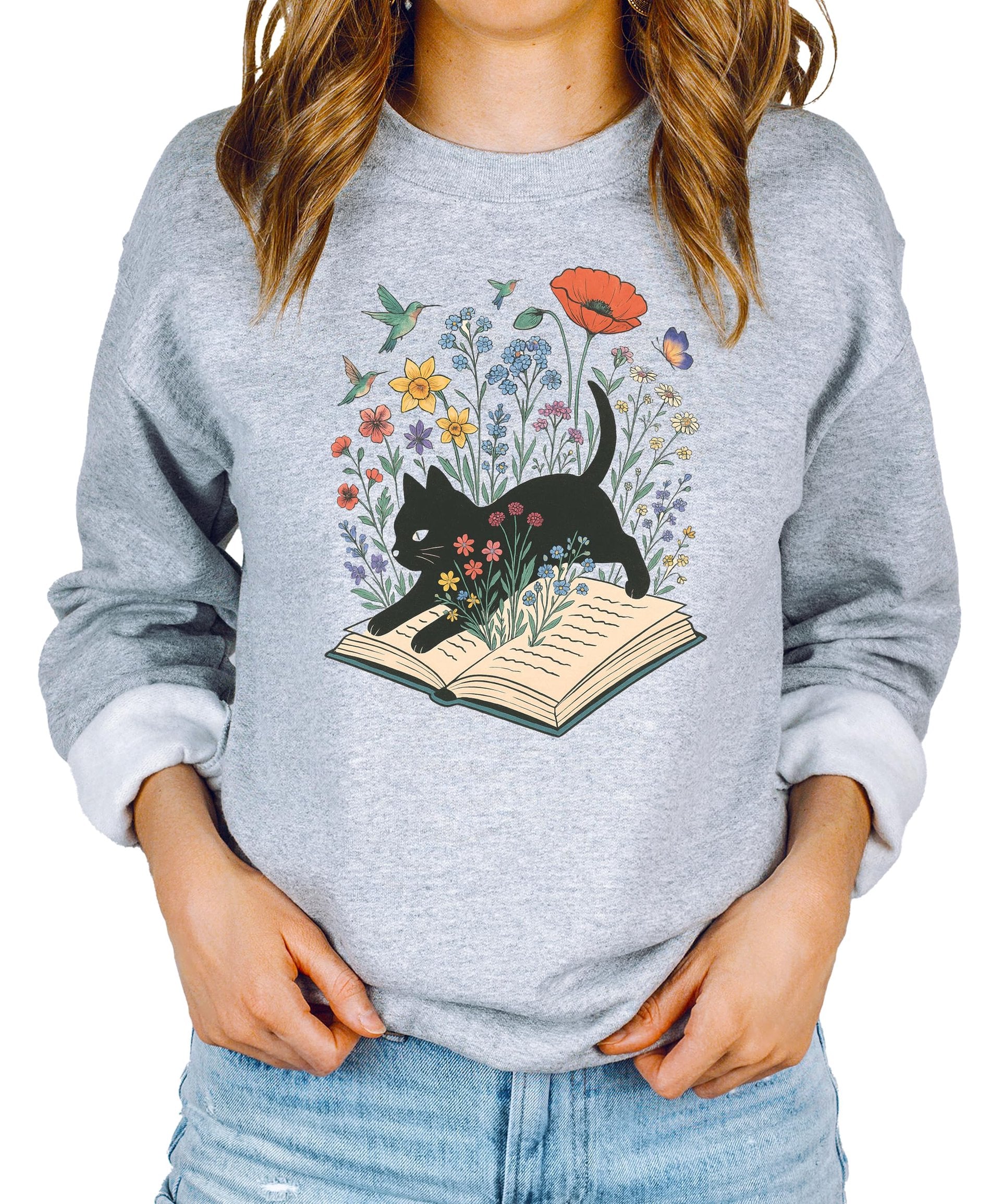White sweatshirt with black cat sitting on open book surrounded by colorful flowers, birds, and butterflies — bookworm graphic design