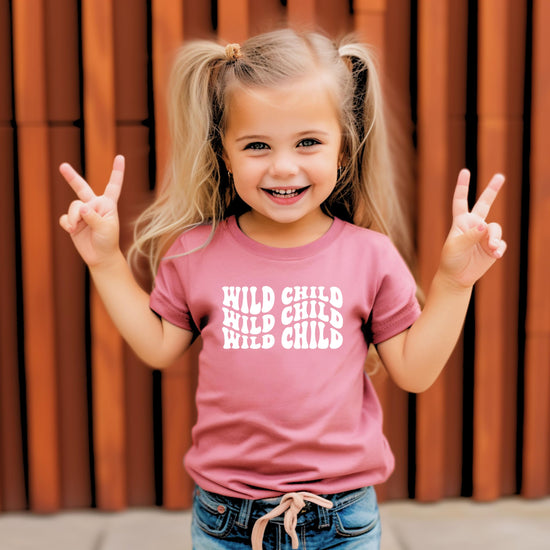 Flat lay of Olive Loves Apple Wild Child peach baby tee with bold white retro graphic