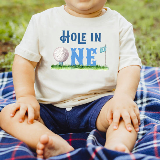 Close-up of Hole in One 1st birthday shirt graphic with golf ball and flag print on natural fabric