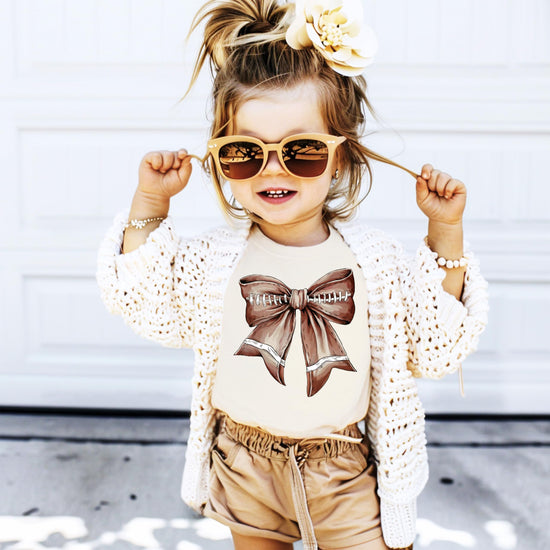 Young girl wearing a football bow shirt in natural color, perfect for game day and fall season