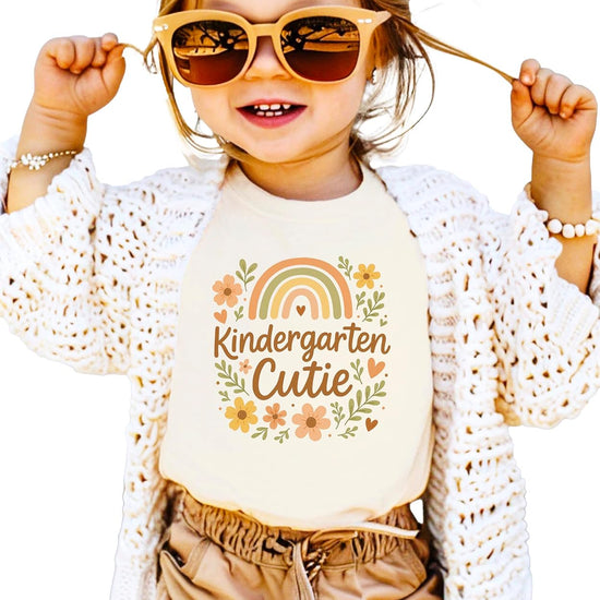 Flat lay of Kindergarten Cutie girls' natural tee showing full rainbow and daisy print detail
