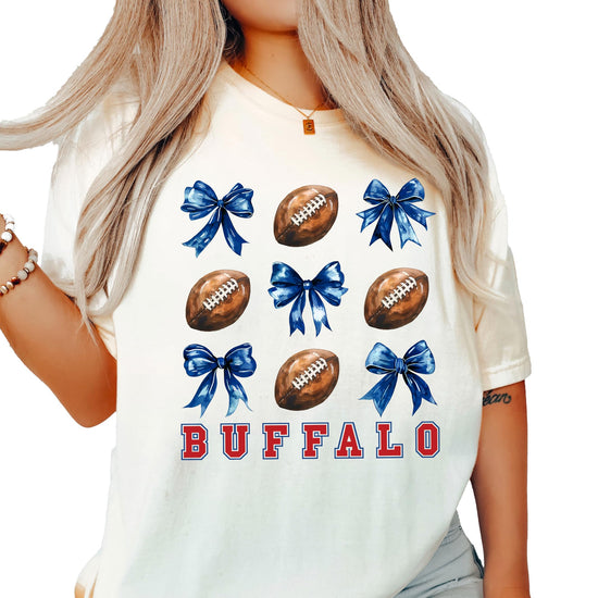 Ivory women's Buffalo football spirit tee with red lettering, footballs, and blue coquette bows graphic