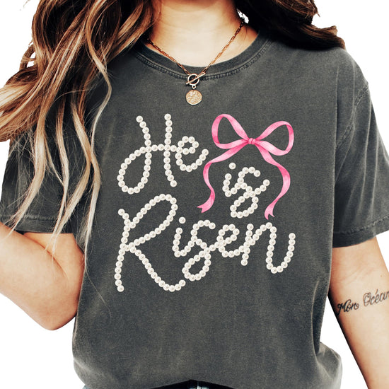 Women's He Is Risen Easter graphic tee with pearl-style lettering and pink coquette bow design