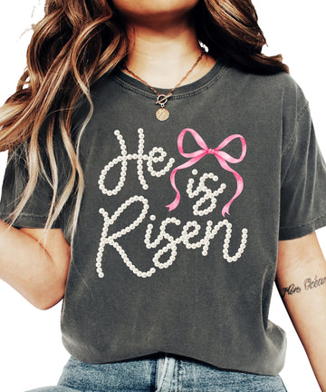 Women's He Is Risen Easter graphic tee with pearl-style lettering and pink coquette bow design