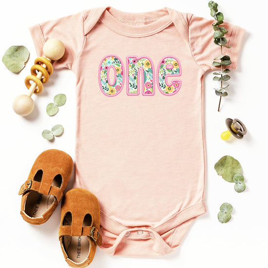 Flat lay of peach and pink baby girl first birthday bodysuit with floral one print