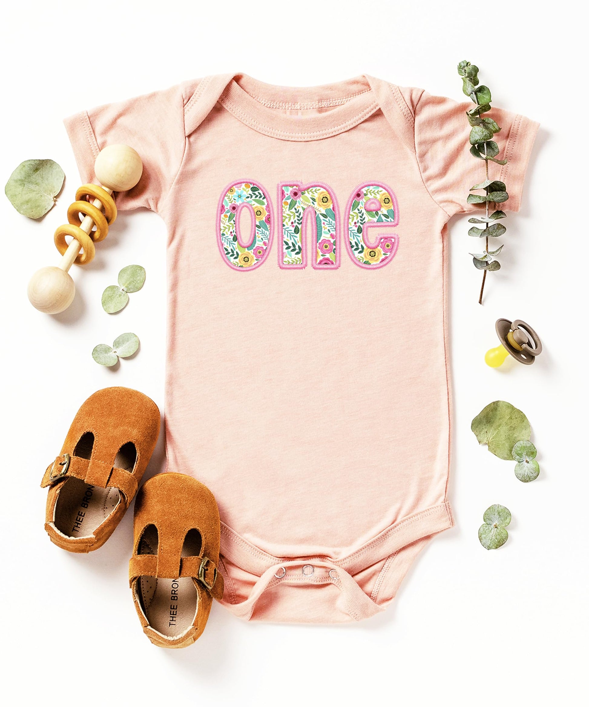 Flat lay of peach and pink baby girl first birthday bodysuit with floral one print