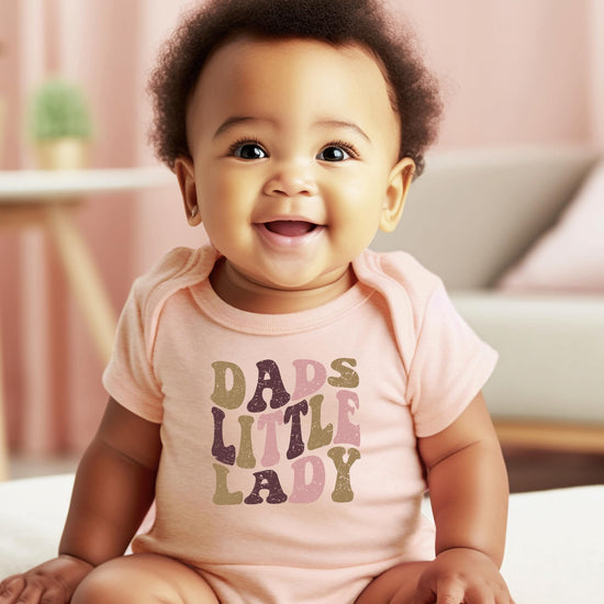 Flat lay of peach snap bodysuit with Dad's Little Lady distressed font design