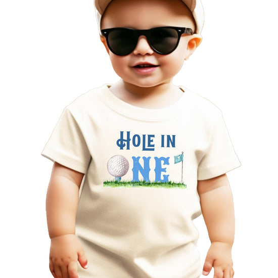 Boys toddler natural tee with Hole in One golf-themed first birthday graphic featuring golf ball, flag, and grass design