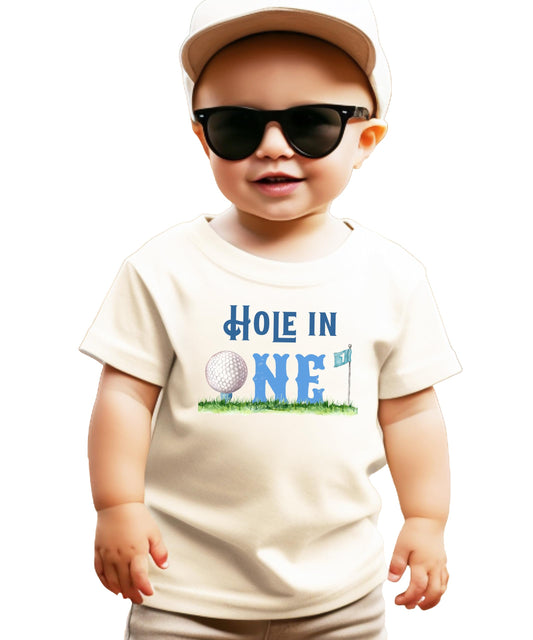 Boys toddler natural tee with Hole in One golf-themed first birthday graphic featuring golf ball, flag, and grass design