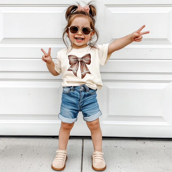 Flat lay of girly football bow t-shirt for toddlers and girls in natural fabric color