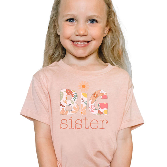 Close-up of floral daisy Big Sister design printed on a soft peach youth tee