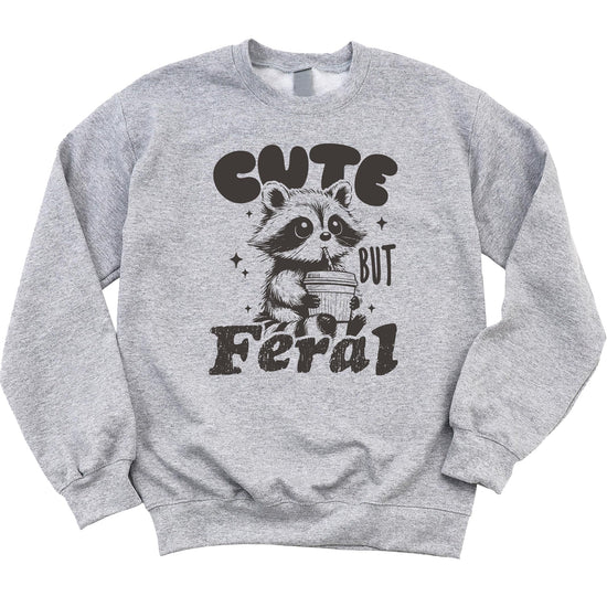Kids pink sweatshirt with a cute raccoon sipping cocoa and the phrase Cute But Feral printed on the front