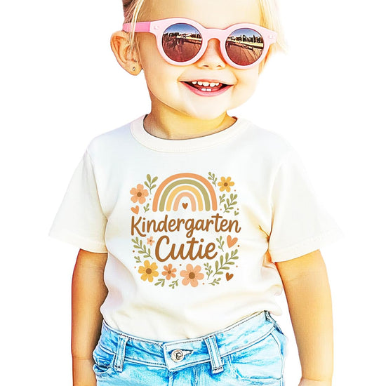 Close-up of hand-lettered Kindergarten Cutie script with wildflower and rainbow design on natural shirt