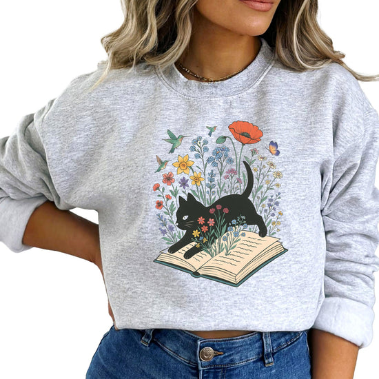 Flat lay of white book lover sweatshirt with black cat nestled in flowers and butterflies graphic print