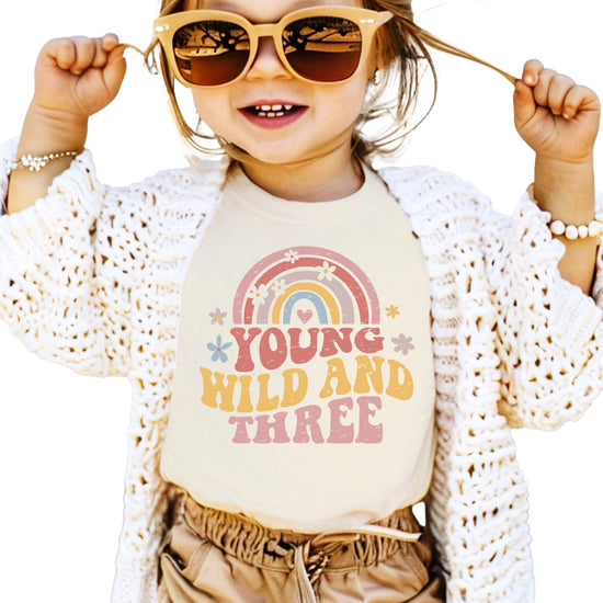 Toddler girl wearing Young Wild Three birthday shirt with retro rainbow graphics