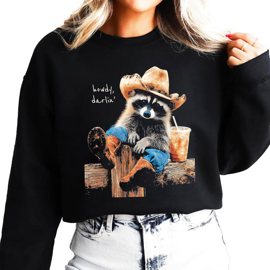 Funny cowboy raccoon crewneck sweatshirt in black with 'Howdy' graphic — raccoon in cowboy hat holding iced coffee