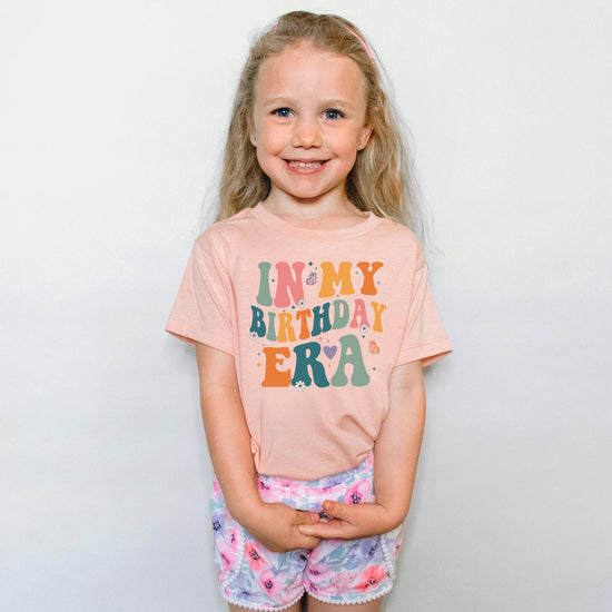 Flat lay of kids birthday graphic tee in peach with bold wavy 'In My Birthday Era' design