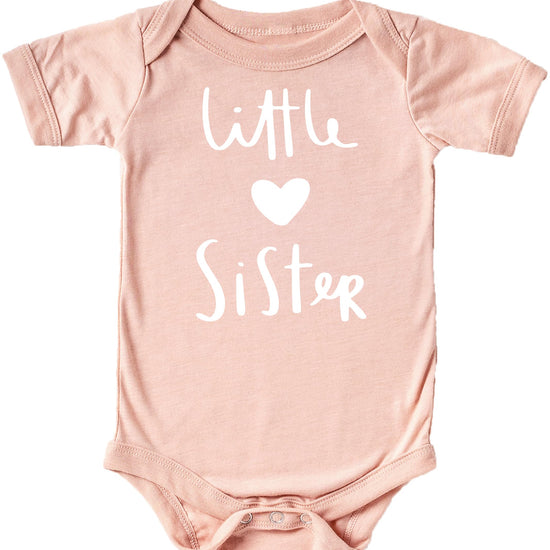 Flat lay of white Little Sister graphic bodysuit with heart detail on mauve surface