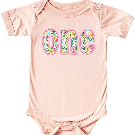 Baby girl wearing a pink floral 'one' first birthday bodysuit with soft ink-printed embroidered-look design