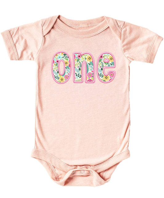 Baby girl wearing a pink floral 'one' first birthday bodysuit with soft ink-printed embroidered-look design