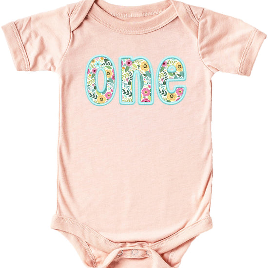 Peach baby girl bodysuit with teal ink-printed floral embroidery celebrating first birthday milestone