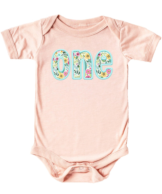 Peach baby girl bodysuit with teal ink-printed floral embroidery celebrating first birthday milestone