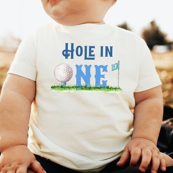 Toddler wearing Hole in One golf-themed first birthday tee, front view on natural colored shirt