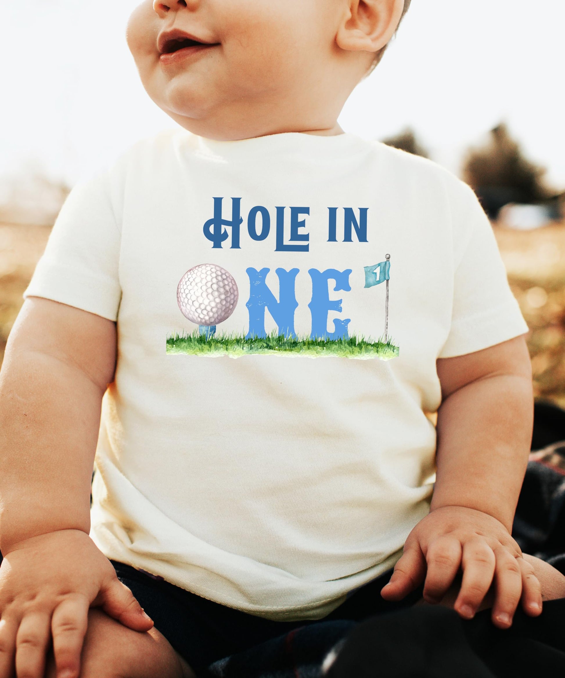 Toddler wearing Hole in One golf-themed first birthday tee, front view on natural colored shirt