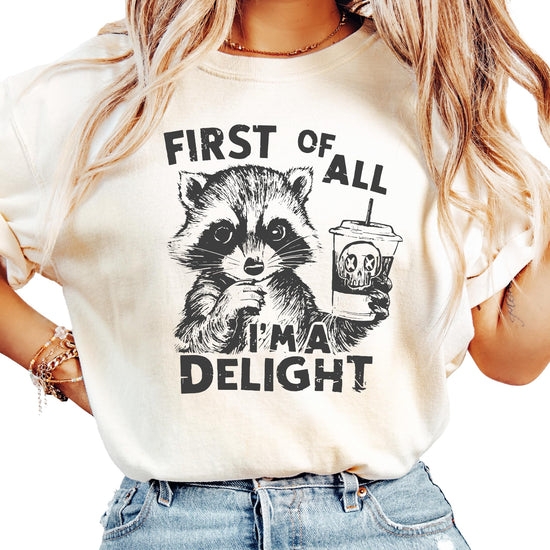 Ivory unisex graphic tee with a raccoon holding a drink and the phrase First of All I'm a Delight