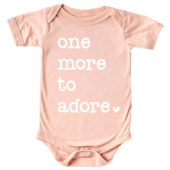White graphic 'One More to Adore' baby bodysuit on military green fabric, newborn pregnancy reveal outfit