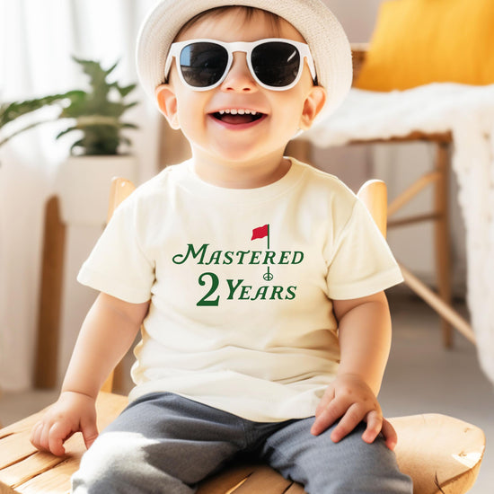 Close-up of golf birthday graphic print on natural kids tee saying Mastered 2 Years