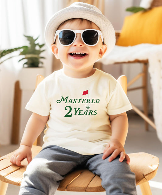 Close-up of golf birthday graphic print on natural kids tee saying Mastered 2 Years