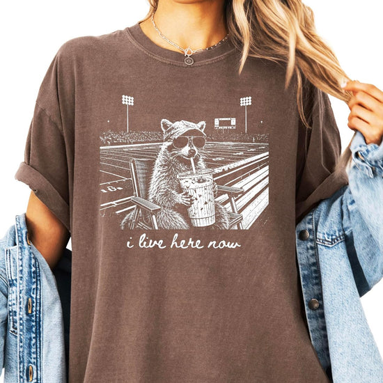 Close-up of raccoon in stadium chair with iced coffee and hand-lettered I Live Here Now script on tee