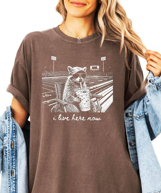 Close-up of raccoon in stadium chair with iced coffee and hand-lettered I Live Here Now script on tee