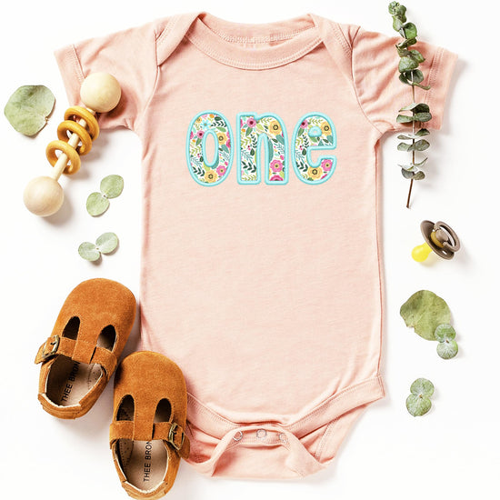 Baby girl wearing peach first birthday bodysuit with teal floral graphic design