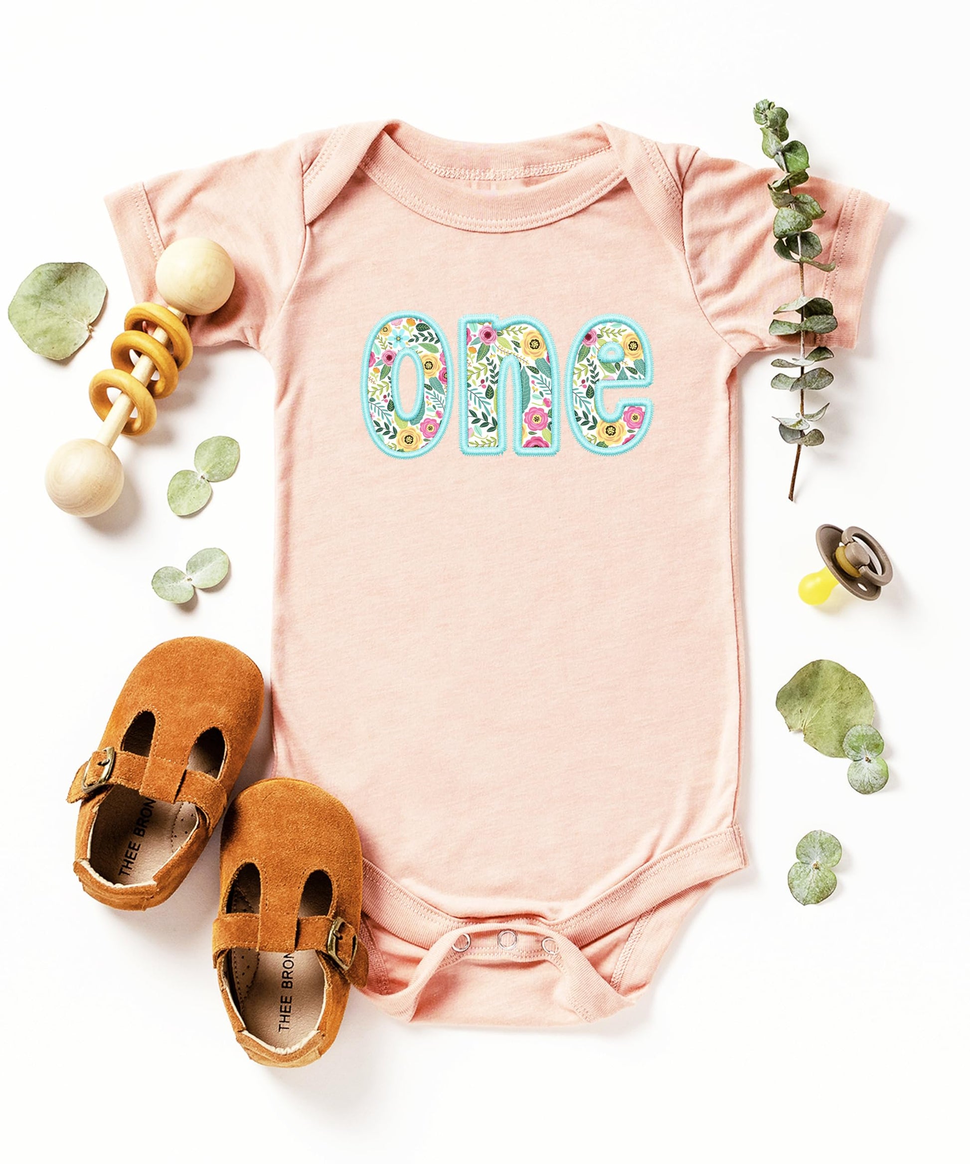 Baby girl wearing peach first birthday bodysuit with teal floral graphic design