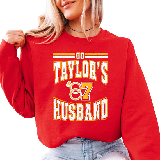 Red crewneck sweatshirt with varsity-style 'Go Taylor's Husband' lettering and wedding rings in red, gold, and white