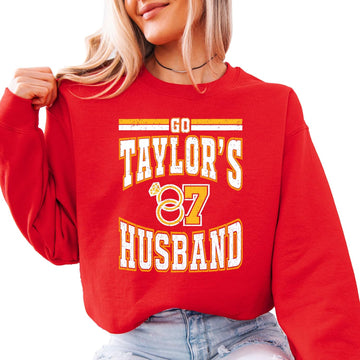 Red crewneck sweatshirt with varsity-style 'Go Taylor's Husband' lettering and wedding rings in red, gold, and white