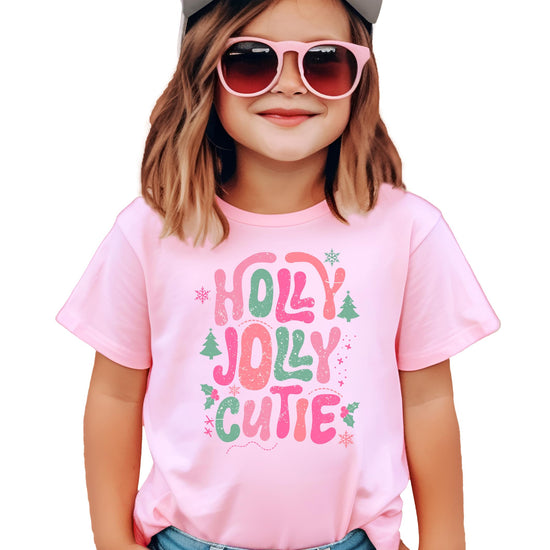 Girls' pink Holly Jolly Cutie Christmas graphic tee with candy cane lettering, Christmas trees, holly leaves, and snowflakes